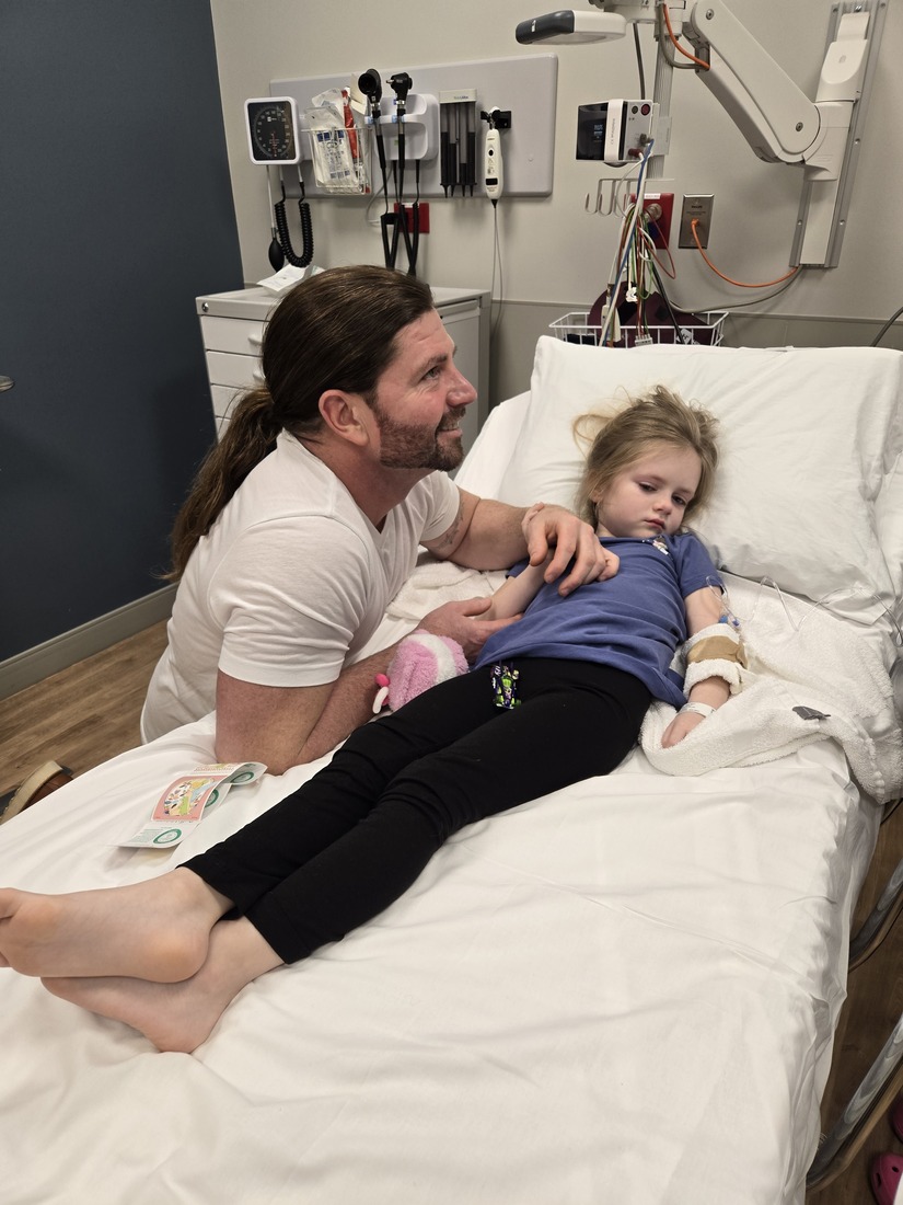 Adam Ironside kneels beside his daughter Iris at St Lukes Marion ER while she receives IV fluids